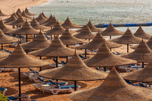View Of A Sandy Beach Covered With Natural Umbrellas. In The Background Is The Sea And Pontoons To Enter The Sea.