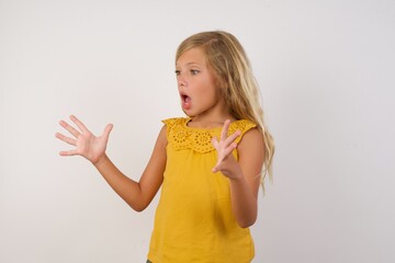 Young blonde kid girl wearing yellow dress over white background shouts loud, keeps eyes opened and hands tense.