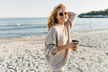 Gorgeous Woman In Sweater Standing At Sea Coast. Fashionable Fair-haired Girl Enjoying Tea Near Ocean