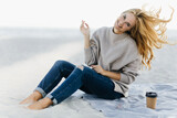 Fototapeta Na drzwi - Positive woman in soft sweater fooling around at beach. Outdoor shot of charming female model sitting on sand with cup of tea