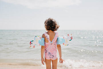 Toddler girl wearing pink swimsuit with unicorns and kids' swimming armbands on the sea