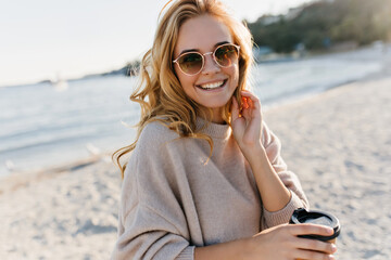 Gorgeous blonde woman in sunglasses and sweater standing at sea coast. Refined curly girl drinking tea at beach and laughing