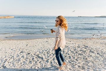 Wall Mural - Charming lady in jeans and sweater looking at sea. Outdoor portrait of adorable girl holding cup of coffee during photoshoot near ocean