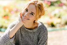 Portrait Of Green-eyed Redhead Girl In High Spirits Looking Into Camera. Woman In Gray Sweater Is Smiling Against Background Of Flowers