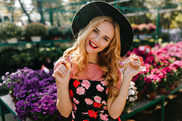 Wall Mural - Inspired female model in hat standing in front of colorful flowers. Portrait of attractive blonde girl relaxing in orangery