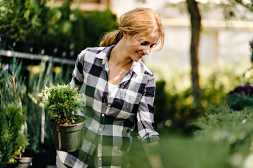 Wall Mural - Spring photo of cute romantic girl in black and white shirt choosing plants. Lady looks at bushes in garden with interest