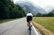 © BublikHaus - Athletic fit cyclist on professional carbon road bike ride on tarmac road towards mountain in distance. Man on bike wears white lightweight jersey and protective helmet. Outdoors sports activity