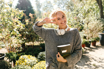 Wall Mural - Portrait of smiling girl in sweater showing sign of peace. Female student posing in park with notebook in her hands