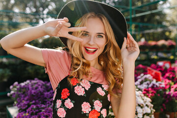 Wall Mural - Excited white girl with blonde hair having fun in greenhouse. Portrait of joyful woman dancing in front of flowers