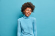 Portrait of glad young Afro American woman smiles broadly, has joyful expression, wears casual blue turtleneck, turns head aside, notices funny scene. Monochrome shot. Happy emotions concept
