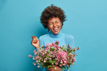Joyful Female Florist Poses With Beautiful Bouquet Of Flowers, Laughs Happily, Has Closed Eyes, Wears Blue Poloneck, Poses Indoor. Afro American Woman Gets Surprise From Husband On Birthday.