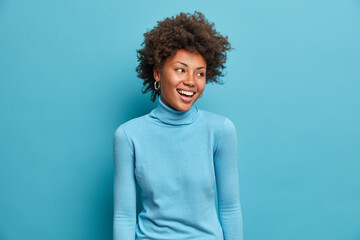 Wall Mural - Portrait of glad young Afro American woman smiles broadly, has joyful expression, wears casual blue turtleneck, turns head aside, notices funny scene. Monochrome shot. Happy emotions concept