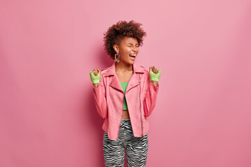 Wall Mural - Overjoyed slim woman has Afro hair cleches fists with joy, feels very happy and lucky after training in gym, motivates to sport wears stylish pink jacket and sport gloves poses indoor exclaims loudly