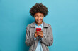© wayhome.studio  - Happy smiling curly haired young woman types message on mobile phone, looks with glad expression at display, wears grey jacket, isolated on blue background. People, lifestyle, technology concept
