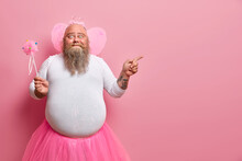 Funny Man Wears Fairy Costume, Invites You On Holiday Or Costume Party, Indicates Right At Blank Space, Holds Magic Wand, Poses Against Rosy Wall. Dad Entertains Children During Birthday Celebration