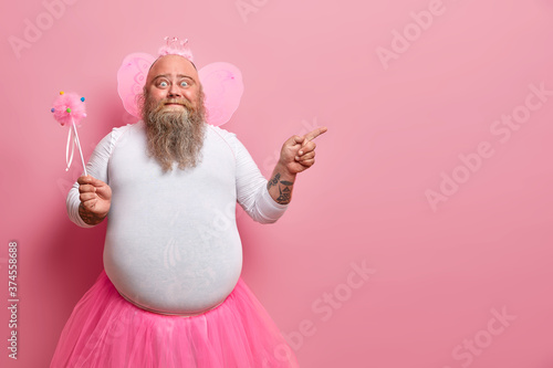 Funny man wears fairy costume, invites you on holiday or costume party, indicates right at blank space, holds magic wand, poses against rosy wall Poster Mural XXL