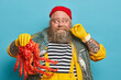 © wayhome.studio  - Cheerful seaman curls mustache, holds big sea creature, dressed in striped jumper and yellow overalls, travels by sea, enjoys summer travel, poses indoor, blue background. Yachting, adventure concept