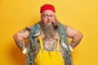 © WHstudio Leushin N - Horizontal shot of plump bearded sailor in overalls keeps hands on hips, thinks deeply while smokes pipe, poses with fishing net, has long beard and tattooed body, isolated on yellow background.