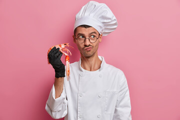 Wall Mural - Professional chef cooks seafood, healthy vegetarian food, holds crayfish, wears white cook uniform, transparent glasses, isolated on pink background, prepares party dinner, poses in restaurant kitchen