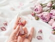 © Konstantin Aksenov - Three graceful bottles for perfume or essential oil on white crumpled fabric. Woman holds pink glass bottle with eastern ornament. Pink rose bouquet and petals as decoration.