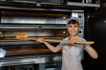 Wall Mural - Young caucasian woman baker is holding a wood peel with fresh bread near an oven at baking manufacture factory.