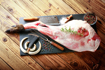 Detail of raw meat, thigh of wild boar on a black plate with rosemary, salt and pepper. the atmosphere of hunting is completed by weapons from wild boar