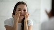 © fizkes - Close up smiling young woman wearing white t-shirt doing facial massage, applying moisturizing cream on under eye skin, looking in mirror, standing in bathroom, enjoying skincare procedure