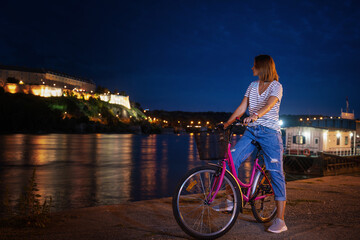 Wall Mural - Young woman with a bike on the Novi Sad embankment overlooking the Petrovaradin fortress, night cityscape travel to Serbia, Balkan countries