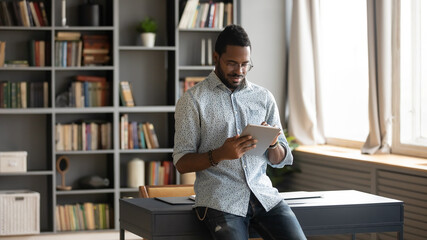 Focused African American man using computer tablet, standing in modern cabinet new work desk, looking at screen, confident young male wearing glasses checking email, working online, writing message