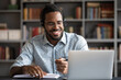 © fizkes - Smiling African American man wearing headphones looking at laptop screen, motivated student writing notes during online lesson, watching webinar, learning language online, sitting at work desk