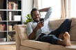 Happy African American man wearing headphones using phone, lying on cozy couch at home, smiling young male enjoying listening to favorite music online, watching video, having fun with smartphone