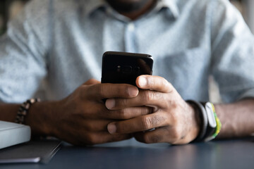 Close up African American man using smartphone, sitting at desk, young male hands holding phone, typing on screen, writing message or browsing apps, searching in internet, chatting online