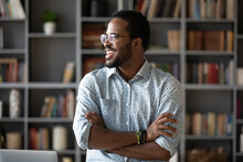 Smiling Positive African American Young Man Wearing Glasses Dreaming About Good Future And Visualizing, New Opportunities, Looking To Aside, Standing With Arms Crossed In Modern Cabinet