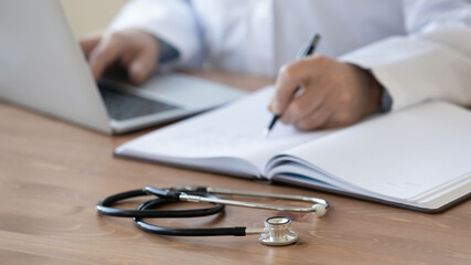 Wall Mural - Close up stethoscope, mature doctor filling documents and using laptop, sitting at work desk in hospital, senior therapist practitioner gp writing illness history, prescription in patient card