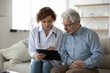 © fizkes - Young woman doctor nurse consulting mature patient during homecare visit, holding clipboard, filling card, illness history, therapist gp giving recommendations to senior man, sitting on couch at home