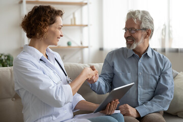 Wall Mural - Smiling mature patient shaking doctor nurse hand, thanking, greeting at meeting, homecare concept, young woman caregiver congratulating senior man with good medical checkup result, using tablet