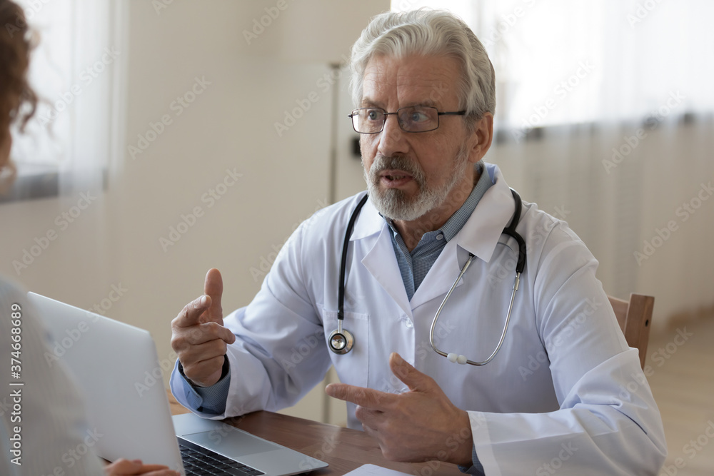 Serious mature doctor consulting female patient at medical appointment, senior therapist gp wearing white uniform with stethoscope sitting at work desk in hospital, discussing treatment results - obrazy, fototapety, plakaty 