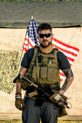 Portrait of serious brutal bearded man in vest with radio device holding riffle against American flag outdoors