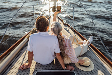 Wall Mural - Rear view of senior man and woman sitting on private yacht and taking selfie with mobile phone