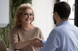 © fizkes - Head shot smiling middle aged 60s businesswoman shaking hands with young male financial advisor, thanking for professional help at office meeting, diverse business people making agreement indoors.