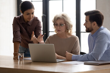 Focused middle aged 60s businesswoman discussing online project presentation with multiracial young colleagues at brainstorming meeting. Diverse employees developing company growth strategy in office.