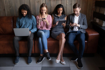 Diverse young people using electronic devices, phones, laptop sitting in row on couch, obsessed by gadgets, African American and Caucasian men and women chatting in social network, playing games