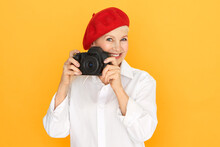 People, Aging, Retirement And Creative Occupation Concept. Portrait Of Senior Female Photographer In White Blouse And Red Bonnet Holding Full Frame DSLR Camera, Taking Pictures In Studio, Smiling