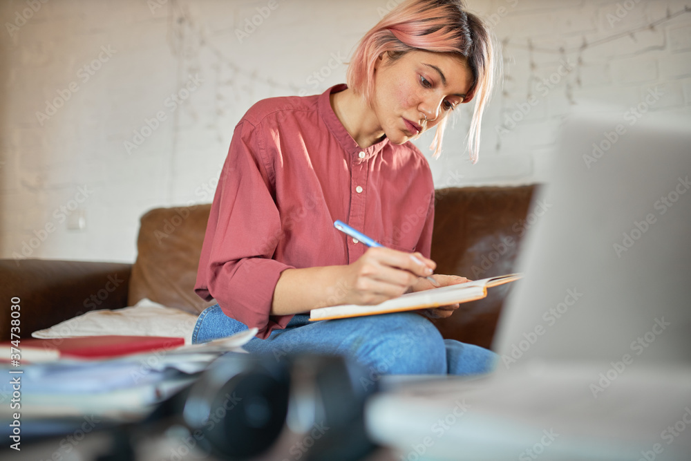 Concentrated young woman copywriter with pink hair working from home making notes in copybook. Cute student girl putting down ideas, writing essay, having focused facial expression, sitting on couch - obrazy, fototapety, plakaty 