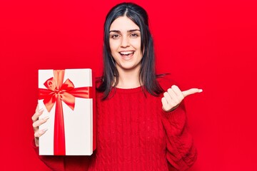 Young beautiful girl holding gift pointing thumb up to the side smiling happy with open mouth