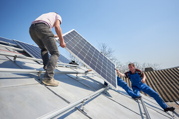 Wall Mural - Male workers installing stand-alone solar photovoltaic panel system. Electricians lifting blue solar module on roof of modern house.