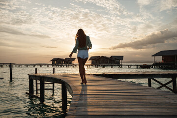 Back view woman  walks over a wooden jetty towards a tropical island during sunset. Luxury travel vacation destination