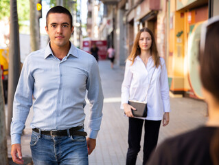 Confident young man dressed in blue shirt and jeans walking through modern city streets on sunny spring day.