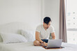 © Sevendeman - Asian man is sitting on the bed and working on his laptop.