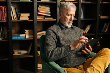 Close-up face of bearded gray-haired mature adult male having break with digital tablet, sitting at home on background of bookshelves in cozy living room with authentic aristocratic interior.